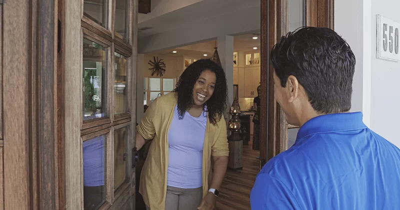 A woman is holding her front door open and smiling at a male Strada Services employee in a blue polo shirt.