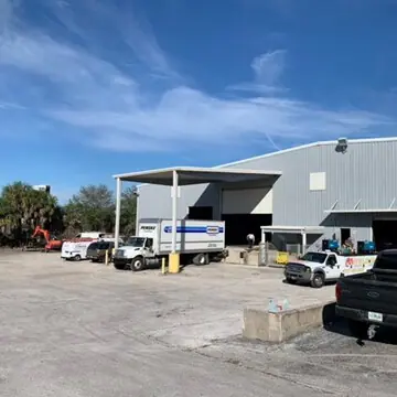 A side view of a warehouse building. Pickup trucks and delivery trucks are parked in front of the building.